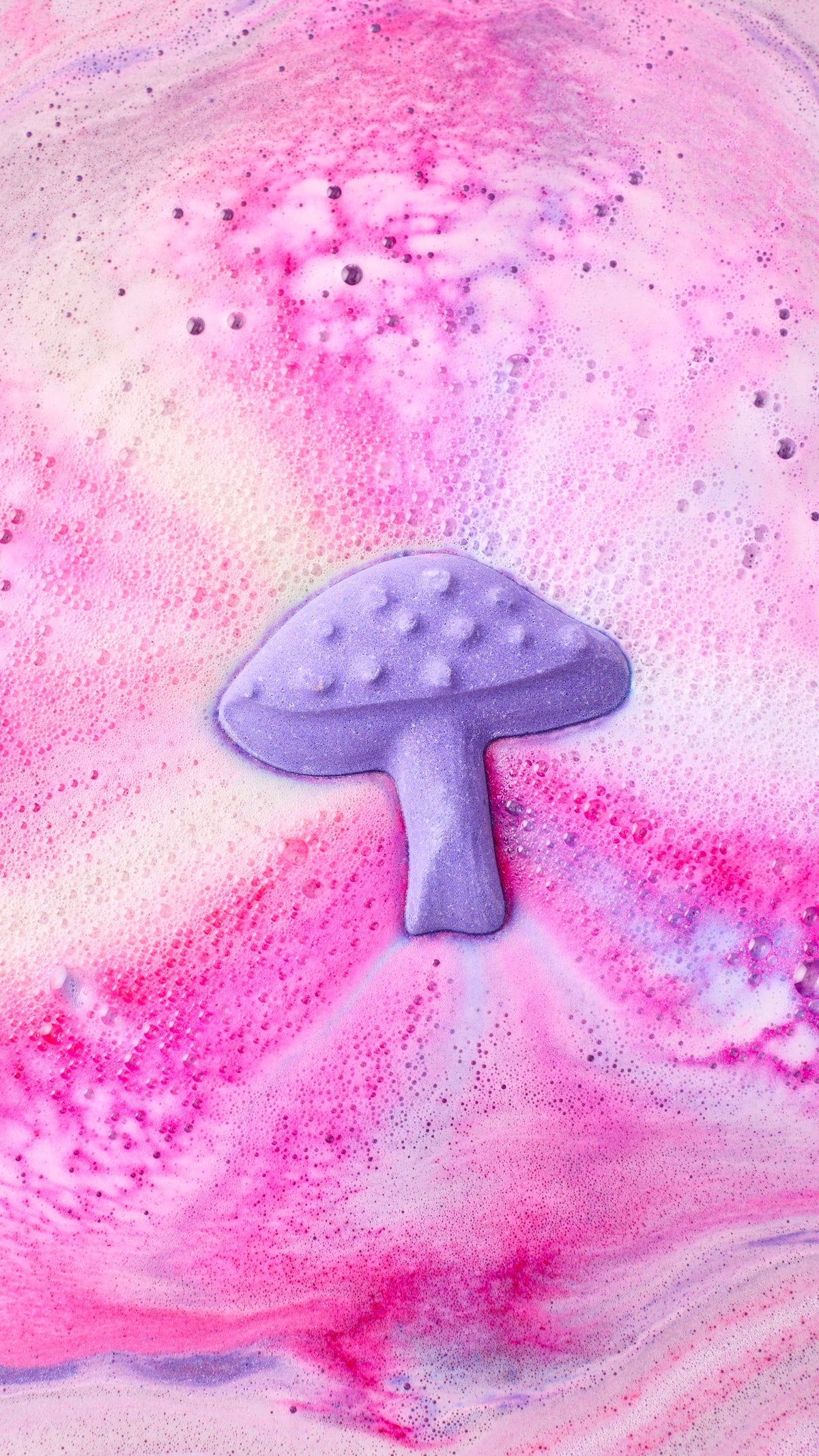An aerial view shows the "Magik Mushroom bath bomb nearly fully dissolved in water, creating a swirling galaxy of pink, violet, and blue foam. The patterns spiral outwards, leaving a trail of thick, colourful bubbles across the water's surface.